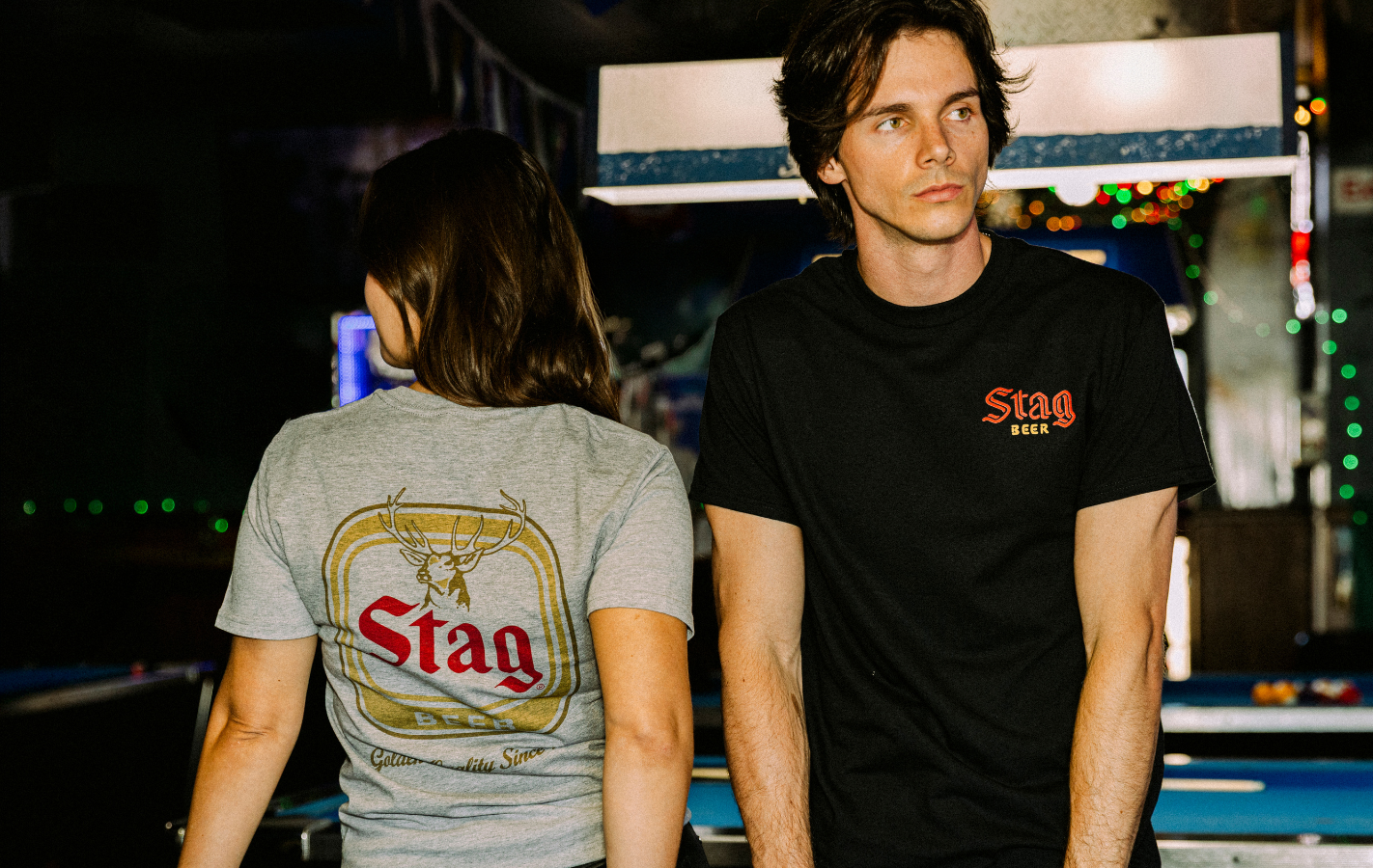 Two people wearing Stag beer shirts standing at a bar counter.